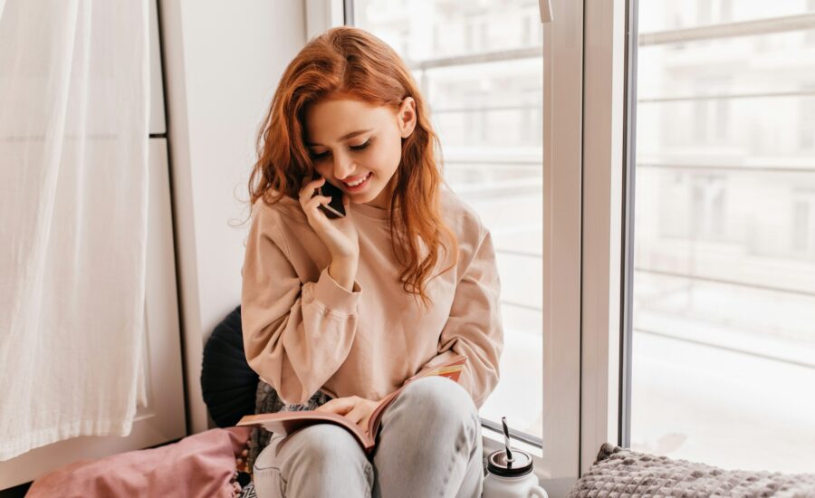 Kobieta w beżowym swetrze siedzi przy oknie z telefonem – lękowy styl przywiązania w związku